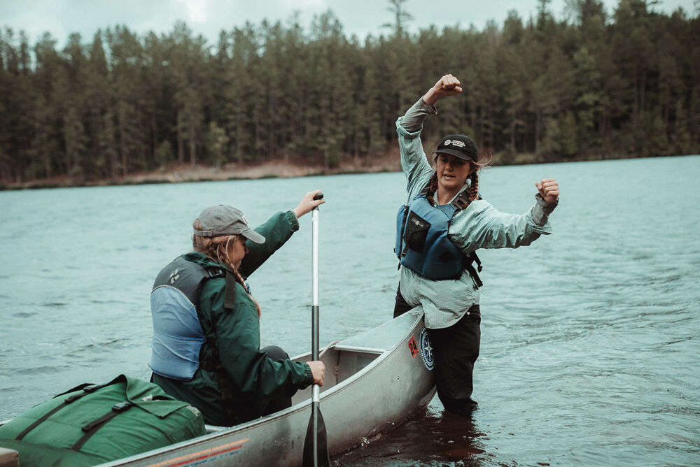Two people are enjoying a canoe trip on a lake surrounded by a forest. One person is seated in the canoe, holding a paddle, while the other stands in the water next to the canoe with their arms raised in the air, seemingly celebrating. Both are wearing life jackets, and a green bag sits in the front of the canoe.